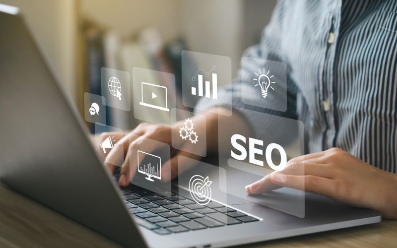 A digital marketer working on a laptop with virtual SEO-related icons hovering above the keyboard, symbolizing different facets of digital marketing such as keywords, analytics, optimization, and global reach.