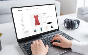 A person using a laptop to browse an e-commerce website showcasing various product categories including women's clothing, with a highlighted product being a red dress priced at $159.00. The website features a clean and modern design with an offer banner displaying a 'Grab deals 50% off' promotion.