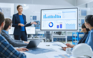 A professional woman in a business suit presents digital marketing analytics on a large screen to colleagues during a meeting. The team, seated at a table with laptops and documents, attentively views the data charts and graphs displayed. The setting is a modern, well-lit office environment with industrial elements.