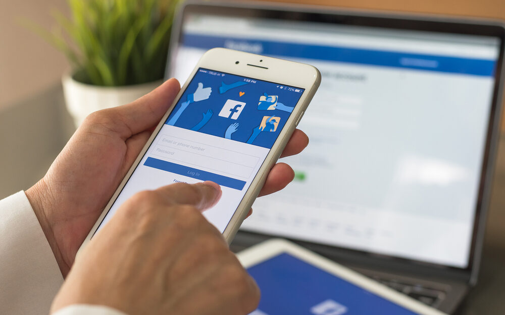 A person logging into Facebook on a smartphone with the Facebook login screen displayed, while a laptop in the background shows the Facebook homepage, representing the use of Facebook ads for business promotion.