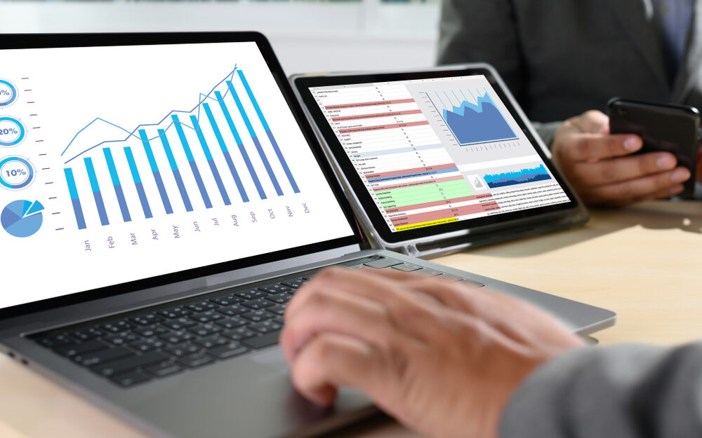 A close-up of a person using a laptop and tablet to analyze data. The laptop screen displays a bar and line graph showing monthly performance metrics, while the tablet screen shows various charts and a data table. In the background, another person is using a smartphone. The setting appears to be a professional office environment, focusing on data analysis and performance tracking.