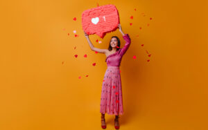 A cheerful woman in a vibrant pink outfit holding a large heart-shaped sign with a "like" icon, symbolizing Instagram engagement, against a bright orange background with heart confetti falling around her.