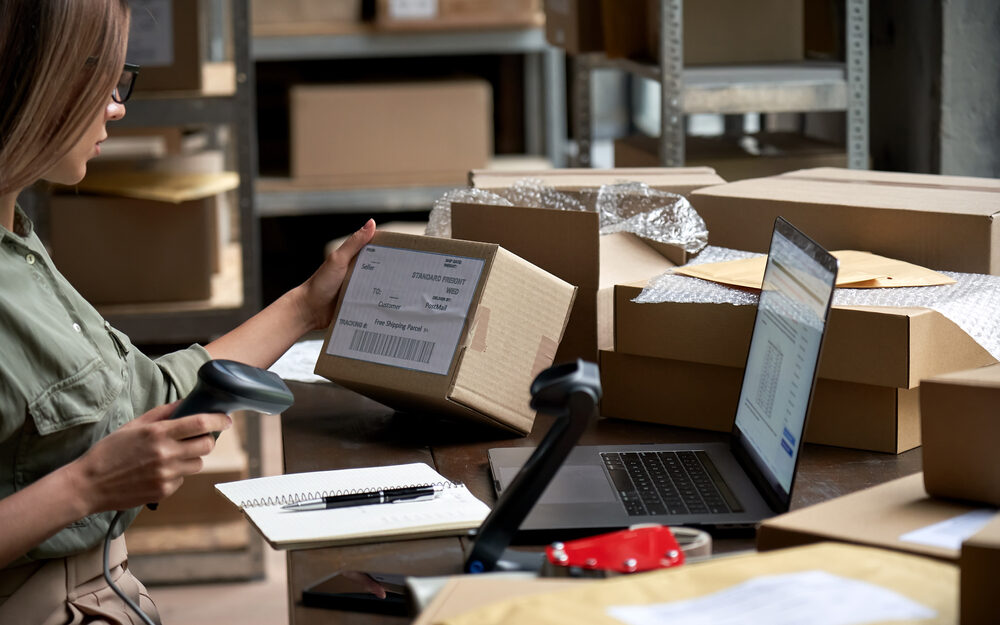 A person working in a warehouse is seen scanning a package label with a barcode scanner. The individual is seated at a desk cluttered with various packages, a laptop, a notebook, and packaging materials. The laptop screen displays a shipping and inventory management system. Shelves filled with more packages and supplies are visible in the background, suggesting a busy environment typical of an e-commerce fulfillment center.