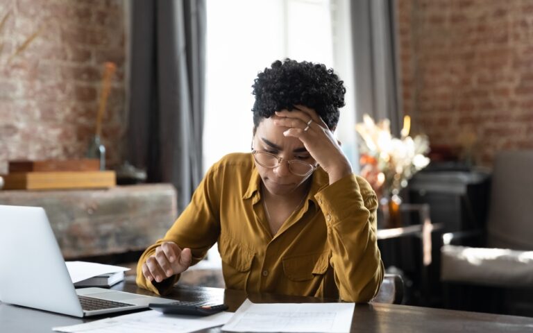 A person sitting at a desk looking stressed while working on a laptop and reviewing documents. The person is wearing glasses and a yellow shirt, with one hand on their forehead, indicating frustration or concentration. The background includes a brick wall, a window with curtains, and a cozy, well-decorated room.